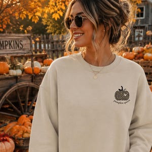 Könnte beinhalten: Ein beigefarbenes Sweatshirt mit Rundhalsausschnitt und einer kleinen schwarzen Grafik auf der linken Brust. Die Grafik zeigt einen Kürbis mit Leopardenmuster und einem kleinen Herz, darunter steht der Text "pumpkin queen" in einfacher Schrift.