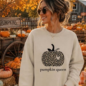 Könnte beinhalten: Ein beigefarbenes Sweatshirt mit Rundhalsausschnitt und einer schwarzen Grafik eines Kürbisses mit Leopardenmuster und einem kleinen Herzausschnitt. Der Text "pumpkin queen" ist in schwarzen Kleinbuchstaben unter der Kürbisgrafik gedruckt.