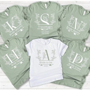 Peut inclure: Plusieurs t-shirts verts sauge et blancs avec des titres de fête de mariage. Chaque chemise présente une grande initiale, des motifs floraux et des phrases telles que "Bride Security", "Bride Squad" et "Bride Crew". Un t-shirt blanc dit "Bride to be 2026."