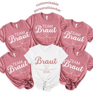 Op de afbeelding: Een verzameling roze en witte t-shirts. De roze shirts zeggen "Team Braut" met een hartontwerp. Het witte shirt zegt "Braut 2026" met een hart. De tekst "customizable personalisierbar" staat bovenaan.