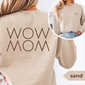 Puede incluir: Sudadera de color arena con el texto "WOW MOM" impreso en marrón en la espalda. Una versión más pequeña del texto está en la parte delantera. La sudadera es de color neutro y tiene cuello redondo.