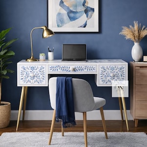 Puede incluir: Un escritorio blanco con diseños florales azules, patas doradas y un portátil. Una silla gris con patas de madera está frente al escritorio. Una bufanda azul está sobre la silla. Una lámpara de escritorio dorada y una pintura abstracta enmarcada también son visibles.