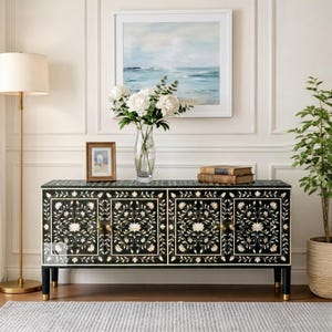 Pode incluir: Um aparador preto com incrustações florais, ferragens douradas e pernas pretas com pontas douradas. O aparador é decorado com um vaso de flores brancas, um quadro emoldurado e uma pintura do oceano. Uma luminária de chão e uma planta em vaso também estão presentes.