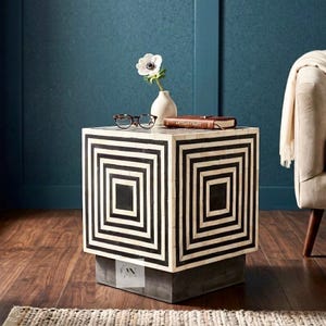 Può includere: Un tavolino a forma di cubo con un motivo geometrico bianco e nero. Il tavolo ha una base grigio scuro ed è sormontato da un piccolo vaso bianco con un fiore, un paio di occhiali e un libro con la scritta "ARTISAN CRAFT".