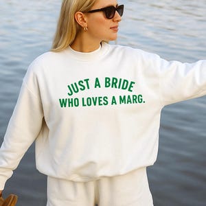 May include: A white sweatshirt and shorts set with the text "JUST A BRIDE WHO LOVES A MARG." in green. The model is wearing sunglasses and holding sandals. The background is a body of water.