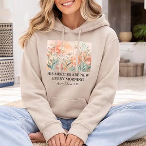 May include: A beige hooded sweatshirt featuring a rectangular graphic of watercolour-style orange and pink flowers. Below the floral design, the text reads: HIS MERCIES ARE NEW EVERY MORNING, followed by the scripture reference Lamentations 3:23.