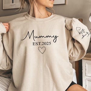 May include: Beige sweatshirt featuring the word "Mummy" in a cursive script, the year "EST.2025", and a heart graphic. The left sleeve displays the names "Freya" and "Lily" with a heart symbol.
