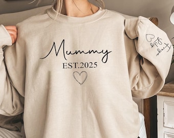 May include: Beige sweatshirt featuring the word "Mummy" in a cursive script, the year "EST.2025", and a heart graphic. The left sleeve displays the names "Freya" and "Lily" with a heart symbol.