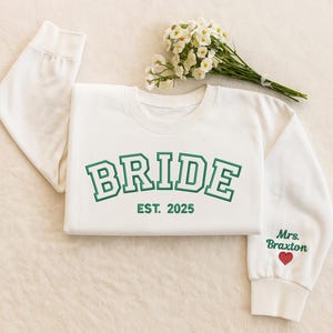 May include: Cream-coloured sweatshirt with the word "BRIDE" in green block letters, with "EST. 2025" underneath. The right sleeve has "Mrs. Braxton" and a red heart embroidered. A bouquet of white flowers rests near the sweatshirt.