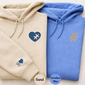 Puede incluir: Dos sudaderas con capucha, una color arena y otra azul Carolina. La sudadera arena tiene un corazón azul con un diseño de pieza de rompecabezas y la letra "S" con un corazón en la manga. La sudadera azul tiene un diseño de pieza de rompecabezas.