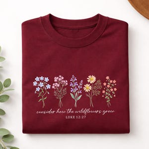 May include: A maroon crewneck sweatshirt featuring a graphic of five distinct wildflower stems in shades of blue, pink, purple, yellow, and orange. Below the flowers, the text reads, "consider how the wildflowers grow" and "LUKE 12:27".