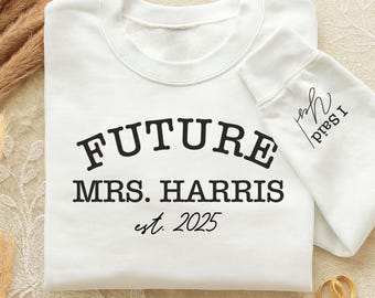 May include: White sweatshirt with the text "FUTURE MRS. HARRIS est. 2025" in black. The sleeve has the text "Yes I Said" in black. Two gold rings are next to the sweatshirt. The word "WHITE" is at the bottom.
