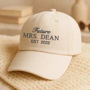 Puede incluir: Gorra de béisbol color crema con el texto "Future MRS. DEAN EST. 2025" bordado en azul marino. La gorra tiene una visera curva y un botón en la parte superior. Está sobre una superficie de punto color crema.