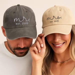 May include: Two baseball caps, one grey and one beige, with the embroidered text "mr. est. 2025" and "mrs. est. 2025". The caps are worn by a man and a woman. The woman is smiling.