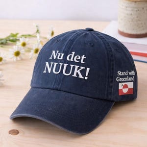 May include: A navy blue baseball cap with the text "Nu det NUUK!" in white. The cap has a curved brim and a small patch with the Greenland flag and the words "Stand with Greenland". A mug and books are in the background.