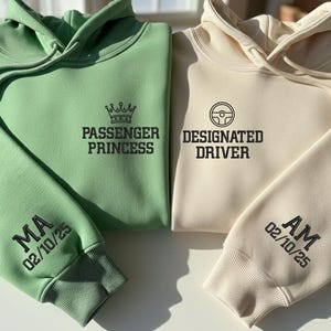 Può includere: Due felpe con cappuccio, una verde chiaro e una color crema. La felpa verde ha "PASSENGER PRINCESS" stampato sul davanti e "MA 02/10/25" sulla manica. La felpa color crema dice "DESIGNATED DRIVER" e "AM 02/10/25" sulla manica.