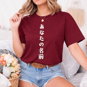 Könnte beinhalten: Burgunderfarbenes T-Shirt mit weißem japanischem Text. Das Shirt ist einfarbig und hat kurze Ärmel. Der Text verläuft vertikal über die Vorderseite des Shirts. Das Model trägt Jeansshorts und einen braunen Gürtel.