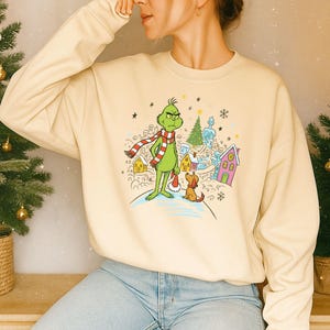 May include: A cream-coloured crewneck sweatshirt featuring a graphic print of a green character wearing a red and white striped scarf, standing next to a brown dog in a snowy village setting with colourful houses, a Christmas tree, and snowflakes.