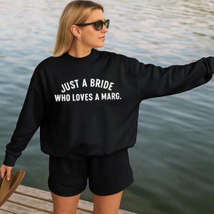 May include: Black sweatshirt and shorts with the text "JUST A BRIDE WHO LOVES A MARG." The person is wearing sunglasses and holding a pair of sandals. The background is a body of water.
