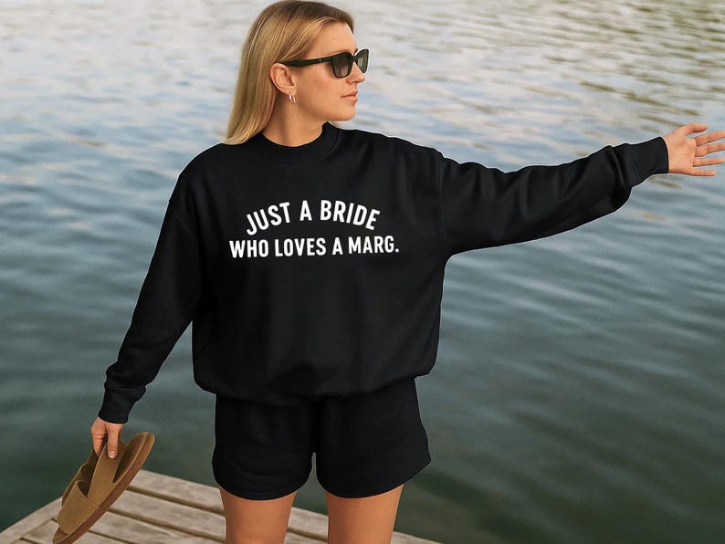 May include: Black sweatshirt and shorts with the text "JUST A BRIDE WHO LOVES A MARG." The person is wearing sunglasses and holding a pair of sandals. The background is a body of water.