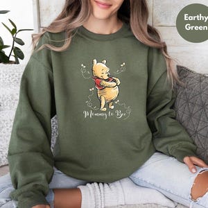 May include: An olive green sweatshirt with a Winnie the Pooh graphic. The cartoon bear is hugging himself, wearing a red scarf, and surrounded by small bees. The text "Mommy to Bee" is written below the bear in a script font.