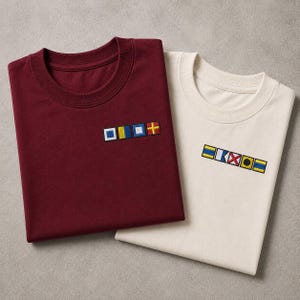 Op de afbeelding: Een bordeauxrood t-shirt en een crèmekleurig t-shirt, elk voorzien van een rij geborduurde nautische signaalvlaggen op de linkerborst. Het bordeauxrode shirt toont vier vierkante vlaggen, terwijl het crèmekleurige shirt zes kleurrijke vlaggen heeft.