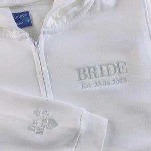 May include: White zip-up hoodie with grey embroidered text: "BRIDE" and "Est 23.06.2023." The left sleeve displays "Dr. & Mrs. P" and a heart. The hoodie has a hood and a small blue tag.