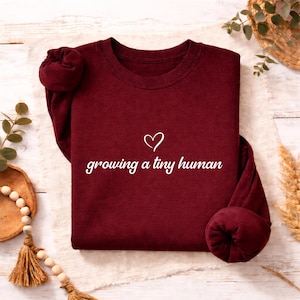 Può includere: Una felpa a maniche lunghe color bordeaux con un'icona a forma di cuore bianco e la scritta bianca "growing a tiny human" stampata al centro del petto. Il capo presenta un girocollo e polsini a coste in tessuto rosso scuro a tinta unita.