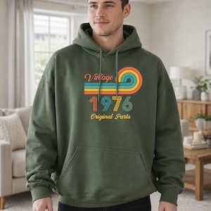 May include: Olive green hoodie with a front pocket and drawstring. The hoodie features a retro design with the words "Vintage 1976 Original Parts" in orange, yellow, blue, and green.