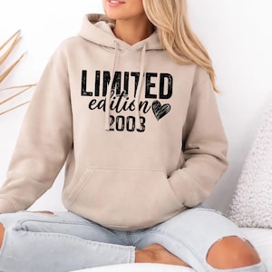 Op de afbeelding: Een beige hoodie met een voorzak en een capuchon met trekkoord. De hoodie heeft de tekst "LIMITED edition 2003" in het zwart, met een hartsymbool. De persoon draagt een lichtblauwe spijkerbroek.