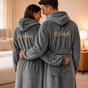 Op de afbeelding: Twee mensen dragen bijpassende grijze badjassen met capuchon. De badjas links heeft de naam "JENNY" in goud geborduurd, terwijl de badjas rechts "Ryleigh" in goud geborduurd heeft. De badjassen hebben een riem en zijn gemaakt van zacht, pluche materiaal.