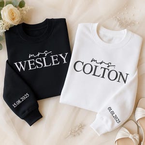 May include: Two crewneck sweatshirts, one black and one white. The black sweatshirt has "mrs. Wesley" printed on the front and "15.08.2025" on the sleeve. The white sweatshirt has "mrs. Colton" and "01.01.2025" on the sleeve.