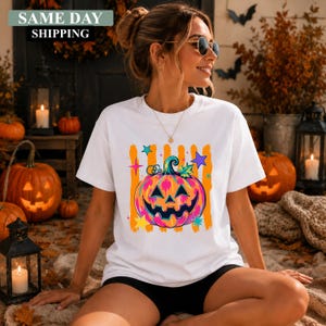 May include: A white short-sleeved t-shirt featuring a graphic of a colourful jack-o'-lantern pumpkin with pink, orange, and blue hues, surrounded by orange vertical stripes and multi-coloured stars. The design is centred on the chest of the shirt.