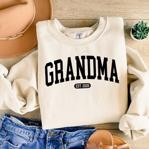 May include: A cream-coloured sweatshirt featuring the word "GRANDMA" in large, black letters. Beneath, the text "EST. 2026" is printed in black. The sweatshirt is styled with blue denim shorts, a brown hat, and sandals.