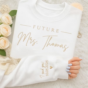 May include: White sweatshirt with gold text reading "FUTURE Mrs. Thomas". The cuff has "I said yes! 10.05.2025" with a heart. A bridal or engagement themed item. The design is embroidered in gold thread.