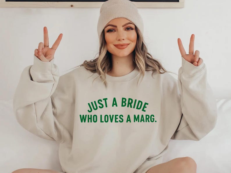 May include: Beige sweatshirt with the text "JUST A BRIDE WHO LOVES A MARG." in green. The person is wearing a beige beanie and making a peace sign with their fingers.