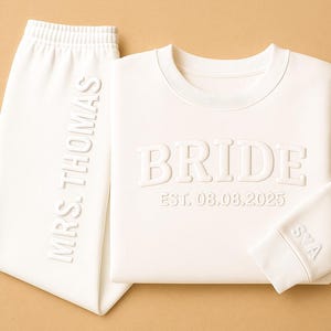 Peut inclure: Un ensemble de survêtement de mariée blanc. Le sweat-shirt porte le mot "BRIDE" et la date "EST. 08.08.2025". Le pantalon de survêtement porte la mention "MRS. THOMAS" verticalement. Le poignet de la manche porte la mention "S♡A".