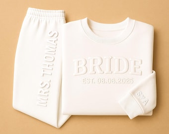 Puede incluir: Un conjunto de chándal de novia blanco. La sudadera lleva la palabra "BRIDE" y la fecha "EST. 08.08.2025". El pantalón de chándal lleva impreso verticalmente "MRS. THOMAS". El puño de la manga lleva impreso "S♡A".