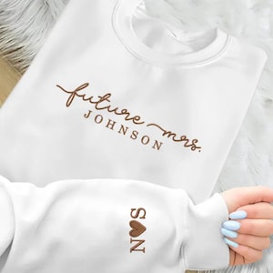 May include: White sweatshirt featuring the text "future mrs. JOHNSON" in brown script. The sleeve shows the initials "S N" with a brown heart. A comfortable top, perfect for a bride.