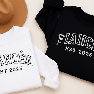 May include: Two sweatshirts, one white and one black, with the word "FIANCÉE" and "FIANCE" in white block letters. Below the word is "EST 2025". A brown hat is in the background. The sweatshirts are likely for a couple.