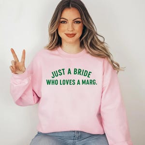 May include: A pale pink sweatshirt featuring the green text: "JUST A BRIDE WHO LOVES A MARG." The crew neck sweatshirt is made of a soft material. The image shows a woman wearing the sweatshirt.