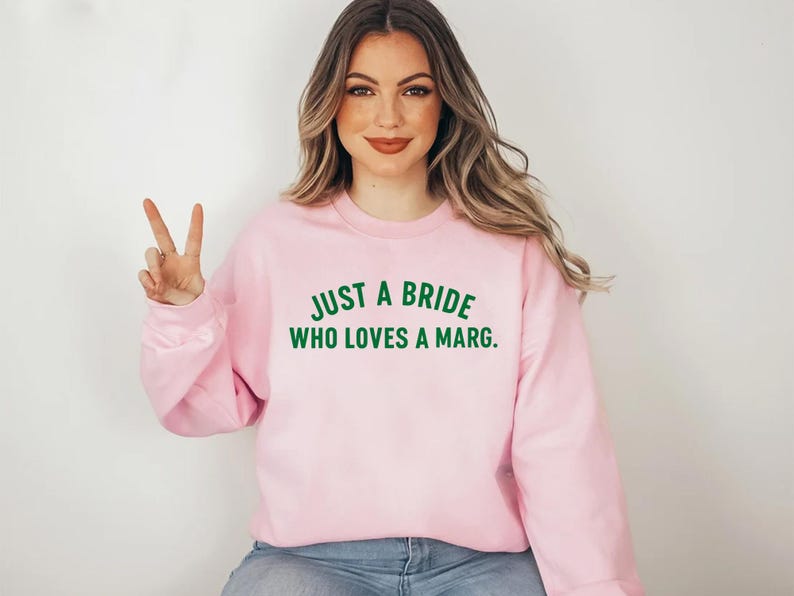 May include: A pale pink sweatshirt featuring the green text: "JUST A BRIDE WHO LOVES A MARG." The crew neck sweatshirt is made of a soft material. The image shows a woman wearing the sweatshirt.
