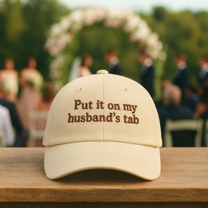 May include: A beige baseball cap with the embroidered phrase "Put it on my husband's tab" in brown lettering. The cap is on a wooden surface, with a blurred wedding scene in the background.