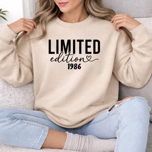 May include: A beige sweatshirt with the words "LIMITED edition" in a bold, black font, with a heart symbol and the year "1986" below. The sweatshirt is worn by a person wearing blue jeans.