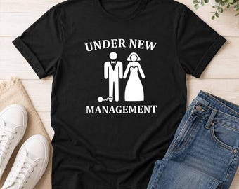 May include: A black short-sleeved t-shirt featuring a white graphic of a stick figure in a tuxedo with a ball and chain on one leg standing next to a stick figure in a wedding dress, with the text "UNDER NEW MANAGEMENT" printed in white.