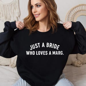 May include: Black long-sleeved sweatshirt with the white text "JUST A BRIDE WHO LOVES A MARG." The sweatshirt is a casual style, suitable for everyday wear. The text is centered on the front.