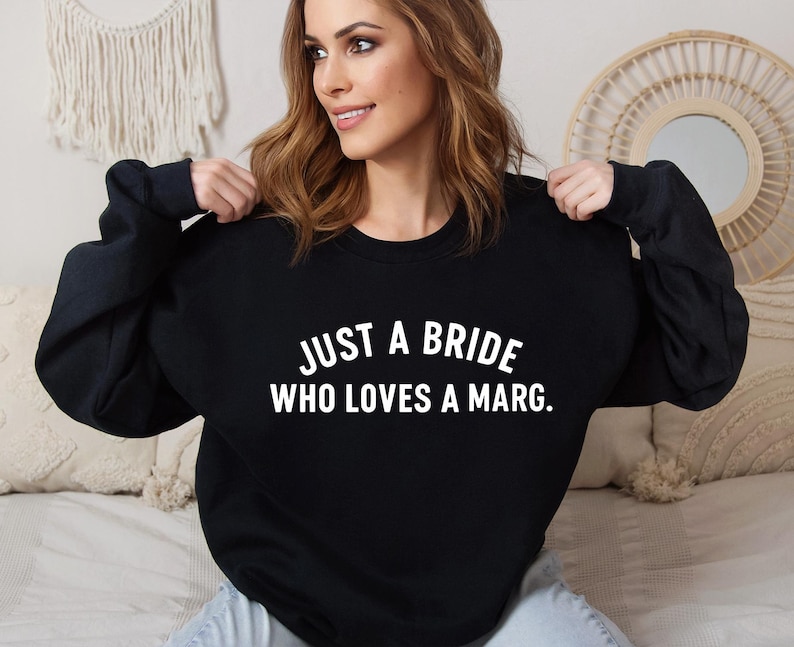 May include: Black long-sleeved sweatshirt with the white text "JUST A BRIDE WHO LOVES A MARG." The sweatshirt is a casual style, suitable for everyday wear. The text is centered on the front.
