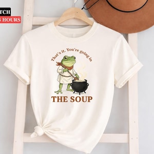 Puede incluir: Camiseta color crema con un dibujo de una rana de dibujos animados con un atuendo de chef, removiendo una olla con la inscripción "THE SOUP". El texto de arriba dice "That's it. You're going in." La camiseta está colgada en una percha de madera.