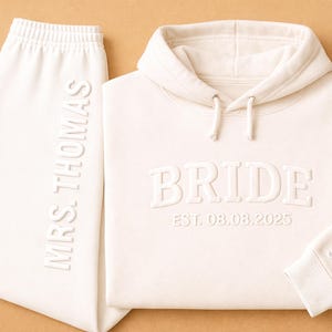 May include: Cream-coloured bridal tracksuit set. The set features a hoodie with the word "BRIDE" and the date "EST. 08.08.2025" embossed on the front, and sweatpants with "MRS. THOMAS" embossed on the side. The hoodie cuff has "S & A" embossed.