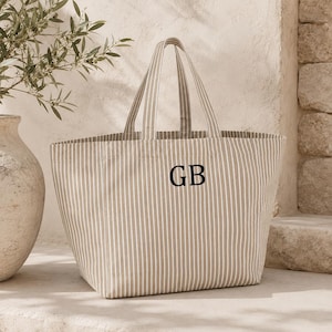 Könnte beinhalten: Eine große Tragetasche mit vertikalem Streifenmuster in Beige und Weiß. Die Tasche hat zwei passende Stoffgriffe und die schwarzen gestickten Buchstaben GB mittig auf der Vorderseite. Das Design ist schlicht mit einer strukturierten Form.